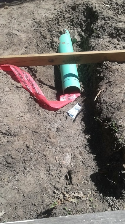 A green plastic pipe is partially buried in a dirt trench, with a red caution tape draped over it. A wooden plank sits across the trench above the pipe, and a small blue and white marker is lying nearby.