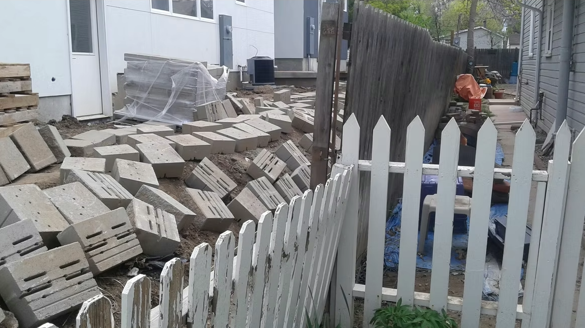 A backyard with scattered concrete blocks, a white picket fence in the foreground, a wooden fence leaning sideways, and construction materials near two houses. The area appears to be under construction or renovation.