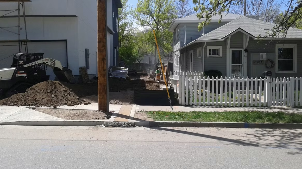 A residential street with construction work in progress between two houses, a dirt pile, a small excavator, a white picket fence, and trees with green leaves in the background.