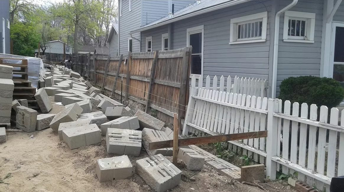 Concrete blocks scattered along a narrow side yard between two houses, with a partially collapsed wooden fence and a white picket fence. The ground is sandy, and trees are visible in the background.