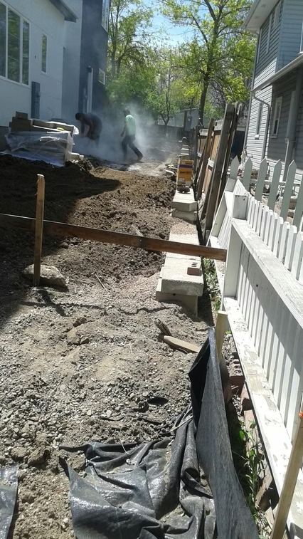 Two workers are visible through dust at a construction site between two houses, with dirt piles, wooden boards, concrete blocks, and a white picket fence along one side under bright daylight.