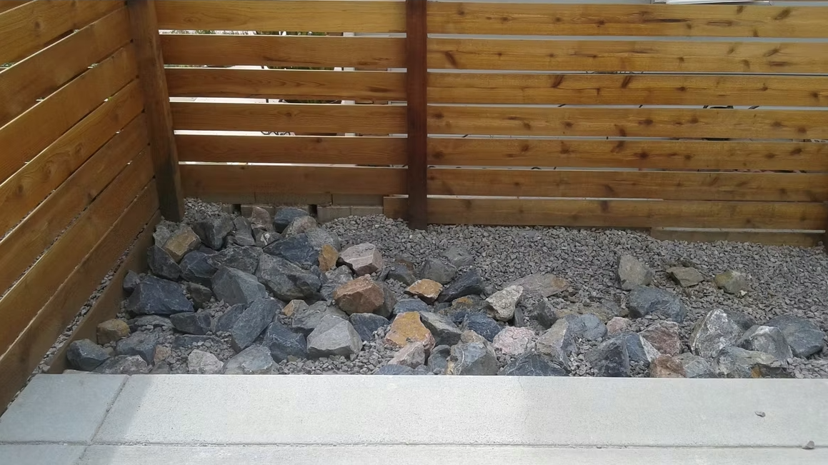 A backyard area with a wooden fence, a section of gray and brown rocks, and small gravel. The foreground includes a concrete or stone edge, likely part of a patio or walkway.