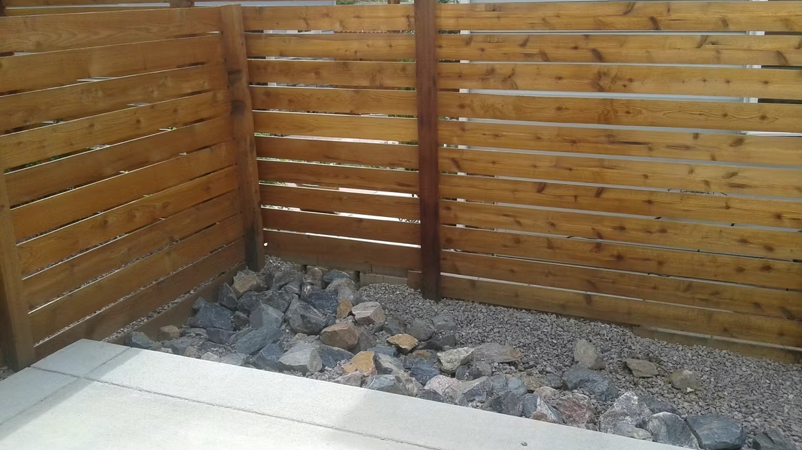 A corner of a yard with a wooden horizontal slat fence, gravel ground, and a pile of assorted rocks along the fence line, next to a concrete patio area.