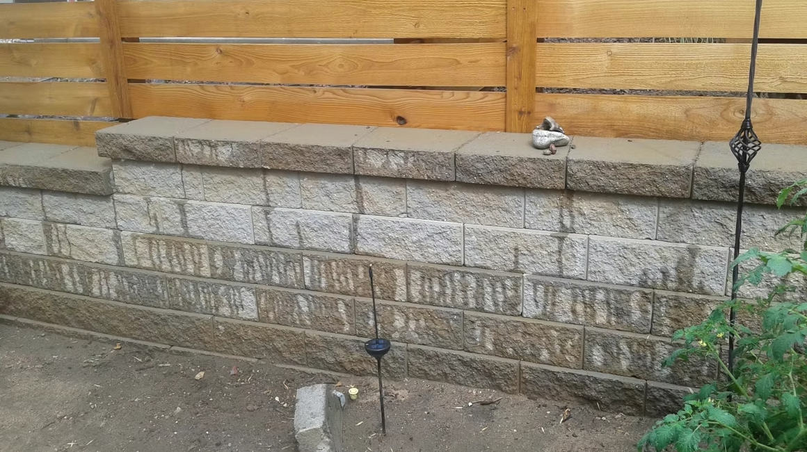 A tiered stone retaining wall with a wooden fence on top, a small garden gnome sitting on the upper ledge, solar garden lights, and bare dirt in the foreground.