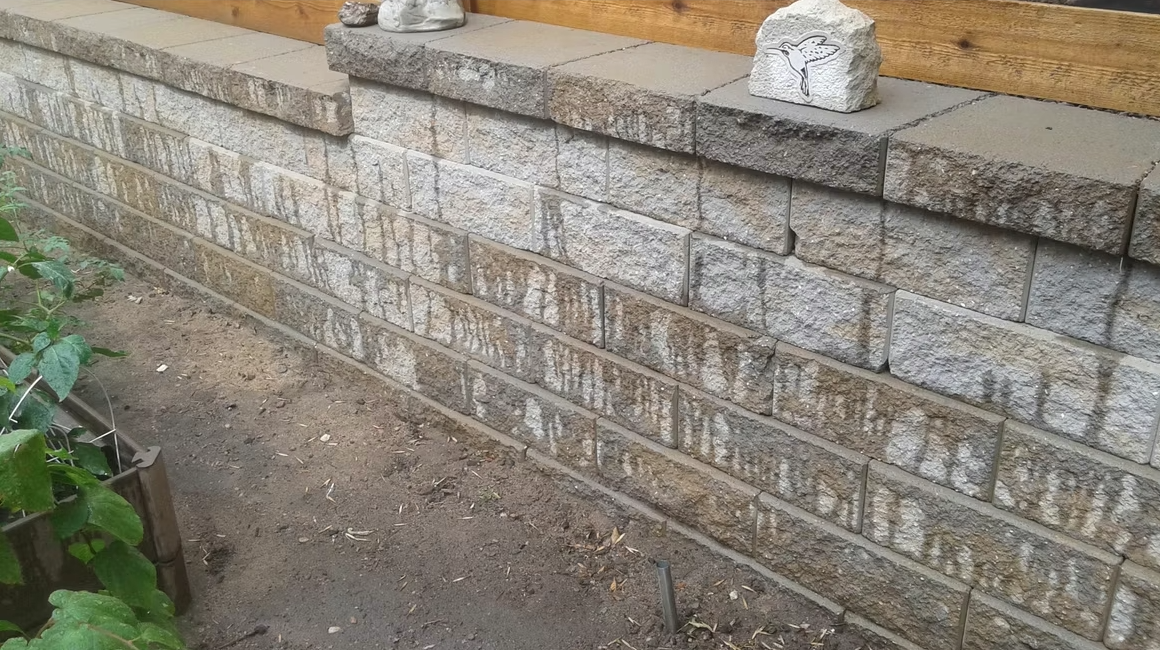 A concrete block retaining wall with visible water stains running down the blocks. There are garden plants to the left, and decorative items, including a stone with a butterfly design, placed on top of the wall.