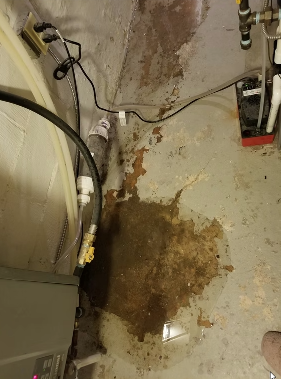 A basement or utility room floor with a large brown water stain and puddle, surrounded by pipes, cables, and equipment near the wall, indicating a possible water leak or flooding issue.