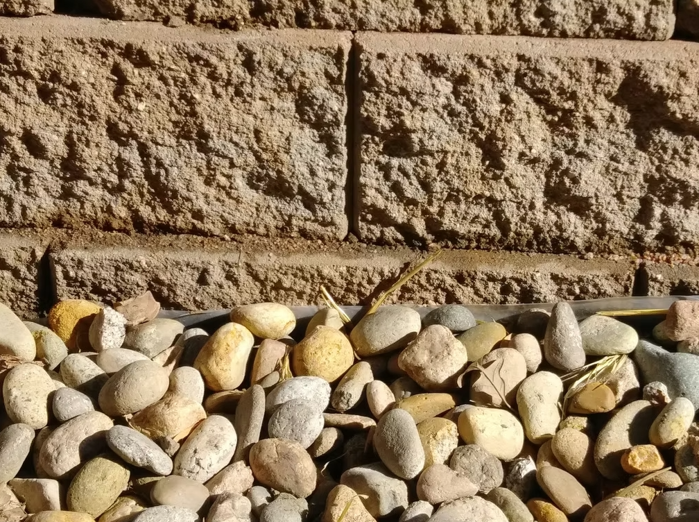A close-up view of smooth, multicolored pebbles in the foreground with a textured brick wall in the background. Sunlight casts shadows, highlighting the rough and uneven surfaces of both the stones and the bricks.