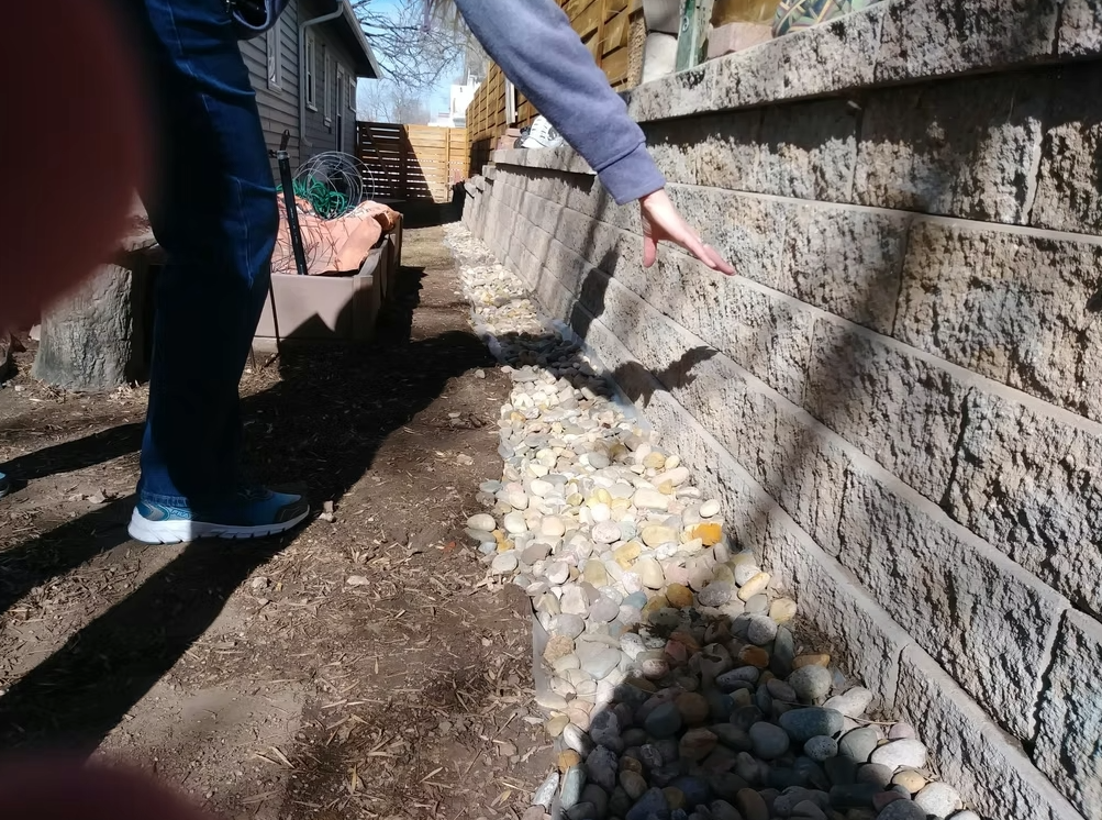 A person stands next to a stone retaining wall, extending their hand toward it. There are light-colored rocks along the base of the wall and soil on the ground. Another persons fingers are visible in the lower left corner.