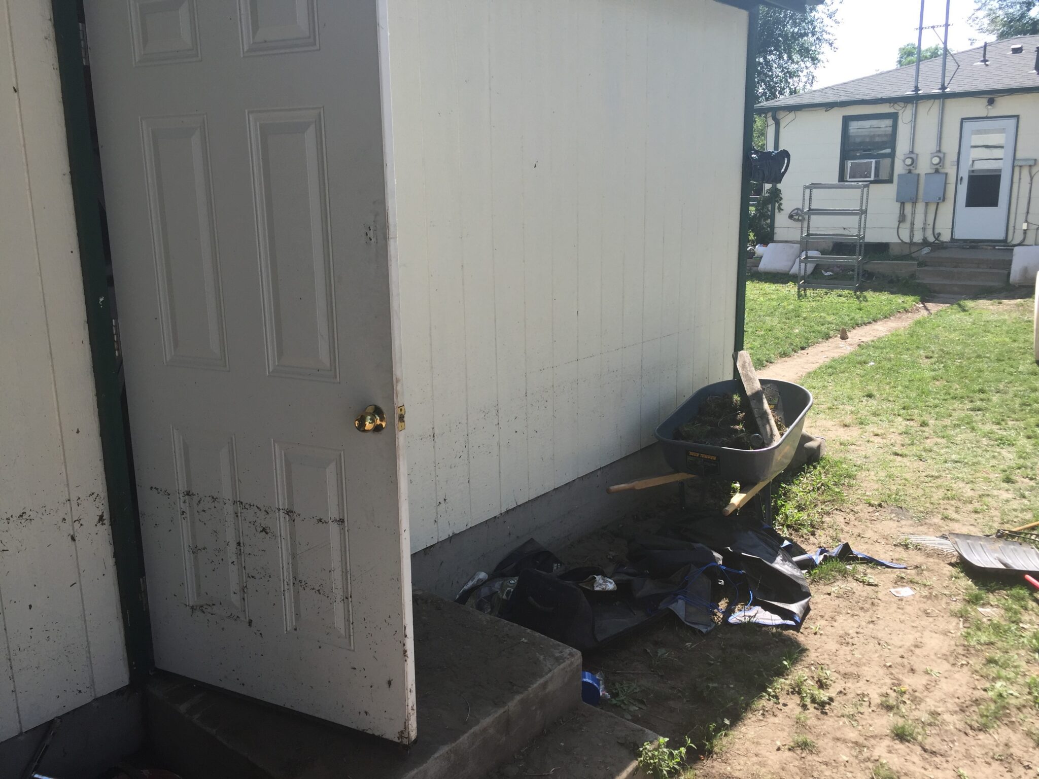A partially open white door on a small white building with a wheelbarrow, shovel, and black bags on the ground nearby. A patchy lawn and another house with utility meters are visible in the background.