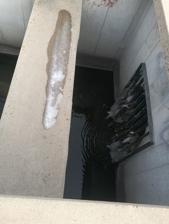 A concrete drainage channel with a partially submerged metal grate, water pooling behind it, and a patch of ice on the upper surface. The walls are grey and the scene suggests cold weather conditions.