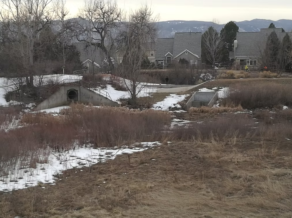 A winter scene with patchy snow on the ground, leafless trees, two concrete tunnels, and houses in the background beneath a cloudy sky and distant mountains.