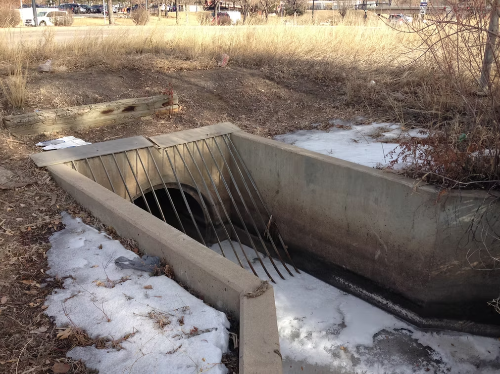 A concrete drainage culvert with metal bars blocking the entrance is partially surrounded by patches of melting snow and dry grass in an outdoor, urban setting.