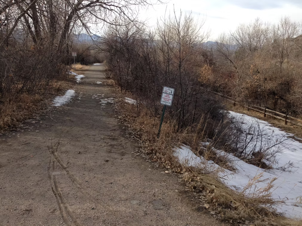 A dirt path lined with leafless trees and patches of snow winds through a wintery landscape. A sign is posted on the side of the path, and a wooden fence runs along a partially frozen creek.