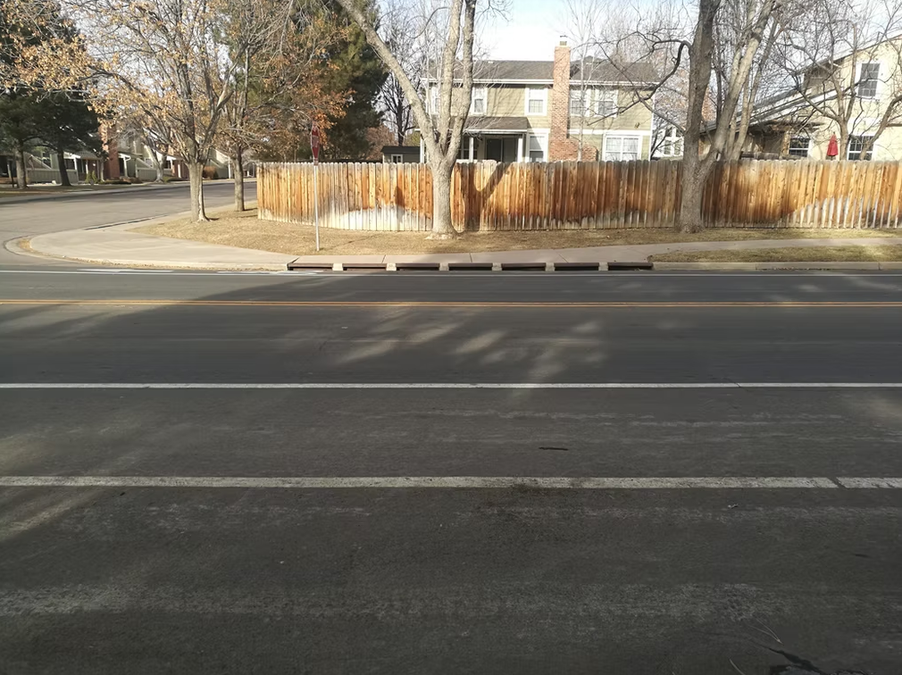 A quiet suburban street with double yellow lines, a wooden fence, trees without leaves, and houses in the background on a sunny day. Shadows from trees are cast across the road.