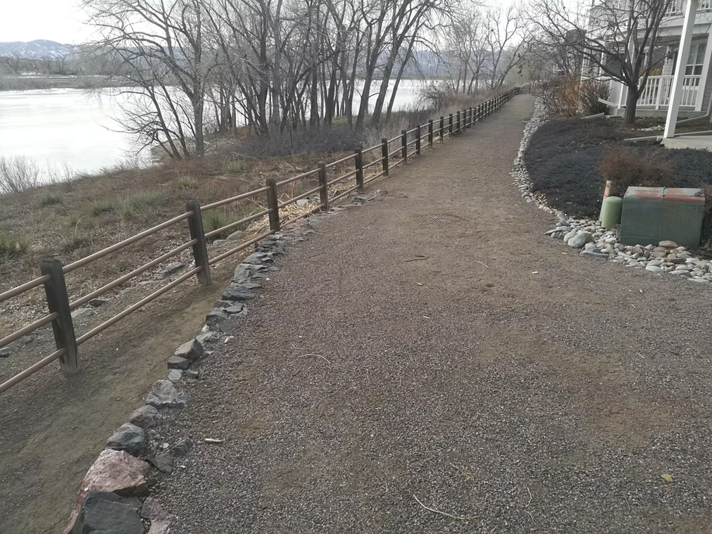 A gravel path bordered by a wooden fence curves alongside a river, with leafless trees and shrubs on the left and houses on the right, under a cloudy sky.
