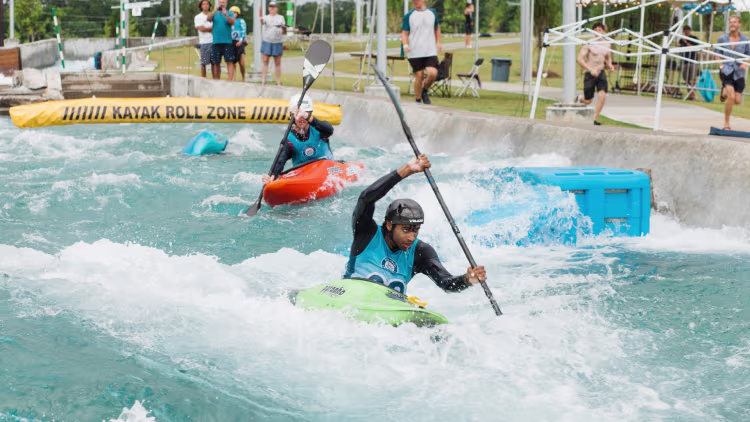 Two people kayaking through a rapid water course under a Kayak Roll Zone banner, with spectators and event staff watching from the sides.