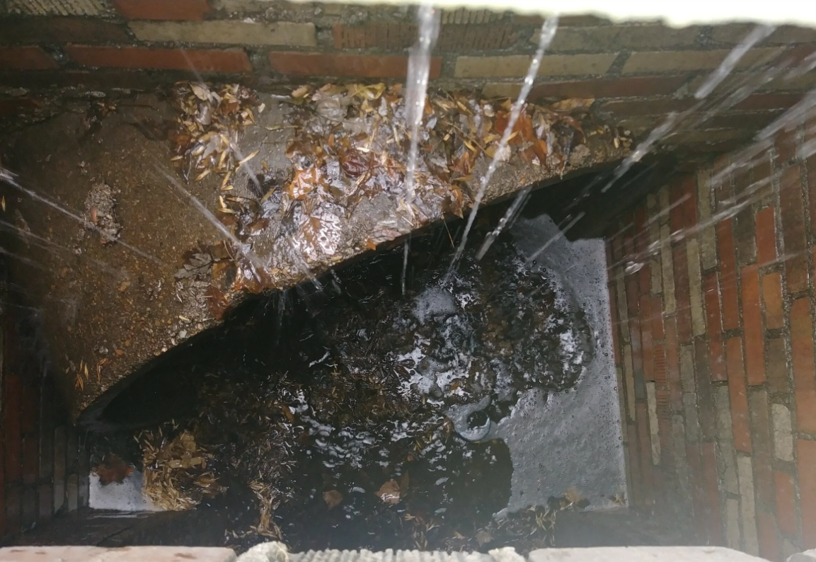 Water streams down into a brick-lined well or pit, with wet leaves and debris scattered on the steps and floor inside, creating a wet and muddy environment.