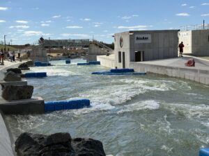 A man-made whitewater rafting course with fast-moving water, blue barriers, rocks, and concrete walls. A few people stand near the structure under a sunny sky with scattered clouds.