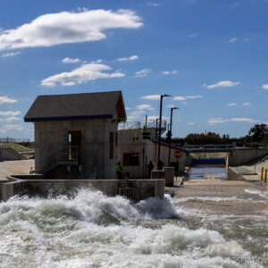 A concrete water facility with a small building sits beside a rushing artificial waterway under a blue sky with scattered clouds; a few people are visible near the structure.