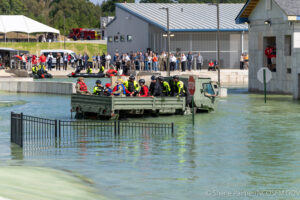 A military vehicle carrying people in safety gear drives through a flooded training area while a crowd watches from behind a fence. A stop sign and partially submerged fences are visible in the water.