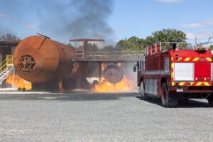 A fire truck approaches a burning aircraft fuselage during a training exercise, with flames and smoke rising around the airplane structure on a sunny day.
